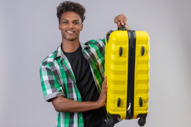 Jeune homme afro-américain voyageur tenant valise souriant joyeusement, positif et heureux