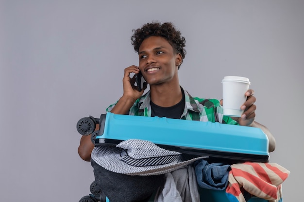 Jeune homme afro-américain voyageur avec valise pleine de vêtements tenant une tasse de café tout en parlant au téléphone mobile souriant avec un visage heureux debout sur fond blanc