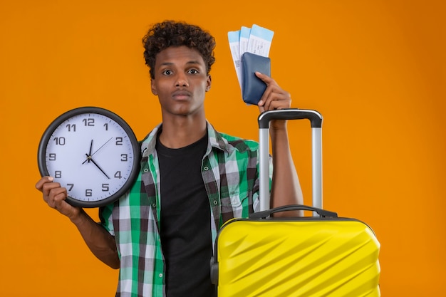 Photo gratuite jeune homme afro-américain voyageur avec valise tenant des billets d'avion et de l'horloge à la recherche d'inquiétude et de confusion