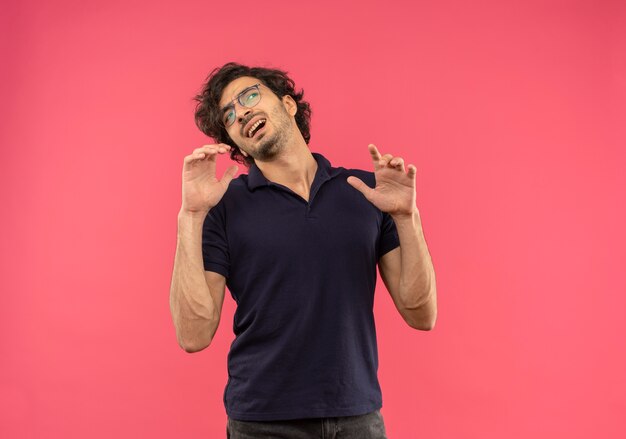 Jeune homme agacé en chemise noire avec des lunettes optiques tient les mains et regarde côté isolé sur mur rose