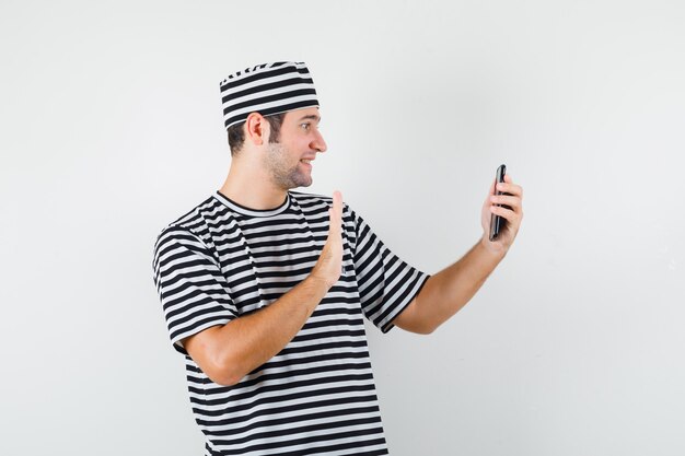 Jeune homme agitant la main sur appel vidéo en t-shirt, chapeau et à la jolly, vue de face.