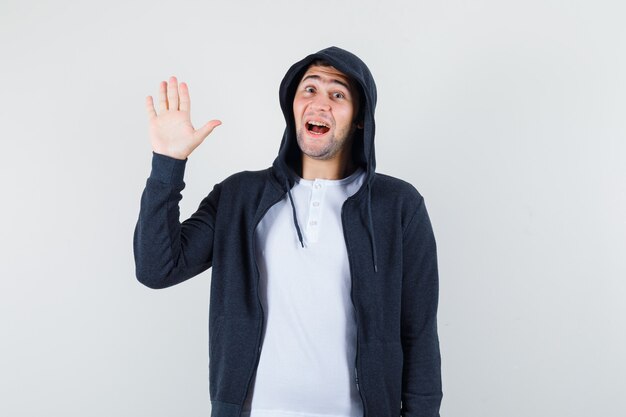 Jeune homme agitant la main pour saluer en t-shirt, veste et à la joyeuse, vue de face.