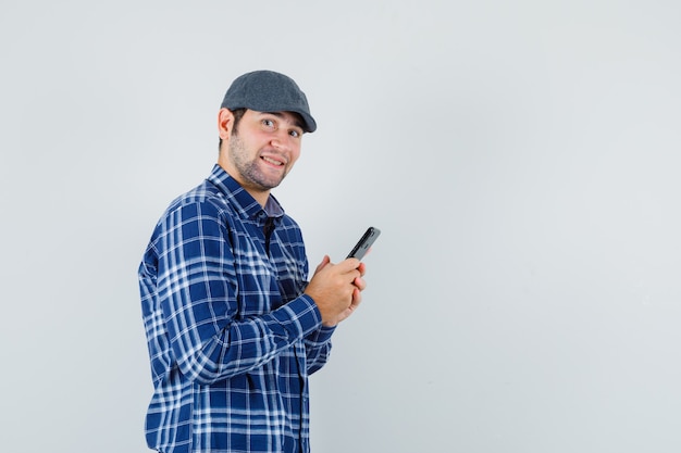 Jeune homme à l'aide de téléphone portable en chemise, casquette et à la joyeuse, vue de face.