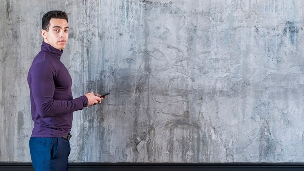 Jeune homme à l&#39;aide de téléphone portable en regardant la caméra debout contre un mur de béton gris