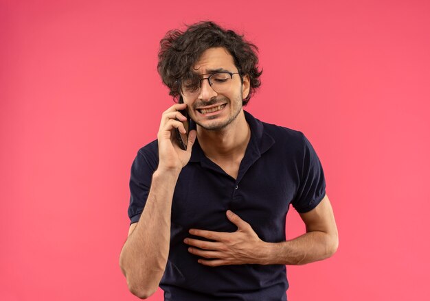 Jeune homme anxieux en chemise noire avec des lunettes optiques parle au téléphone et tient le ventre isolé sur mur rose