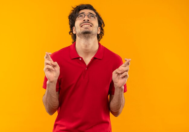 Jeune homme anxieux en chemise rouge avec des lunettes optiques croise les doigts et lève les yeux isolé sur mur orange