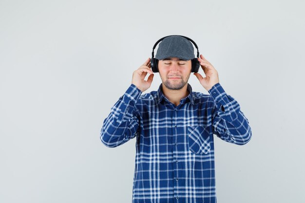 Jeune homme appréciant la musique avec un casque en chemise, casquette et à la recherche de calme vue de face.