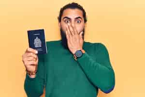 Photo gratuite jeune homme arabe tenant un passeport canadien couvrant la bouche avec la main, choqué et effrayé par erreur. expression surprise