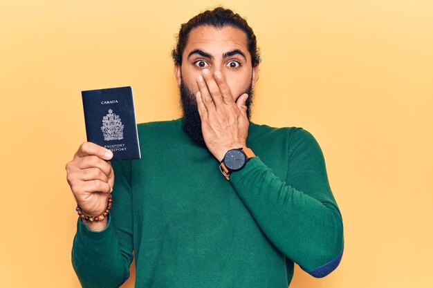 Jeune homme arabe tenant un passeport canadien couvrant la bouche avec la main, choqué et effrayé par erreur. expression surprise