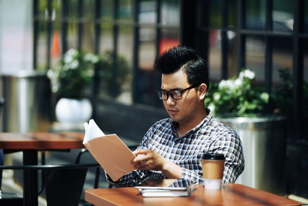 Jeune homme asiatique à lunettes, assis au café en plein air et livre de lecture