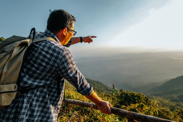 Photo gratuite jeune homme asiatique randonneur avec sac à dos debout et pointer le doigt vers la belle vue sur la vallée au point de vue avec rayon de soleil dans l'espace de copie du coucher du soleil