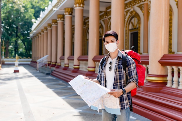 Jeune homme asiatique routard portant un masque facial debout et vérifiant la direction sur une carte papier à la main dans un beau temple thaïlandais,
