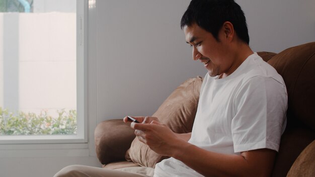 Jeune homme asiatique utilisant un téléphone portable jouant à des jeux vidéo à la télévision dans le salon, un homme se sentant heureux à l’aide de la détente, allongé sur un canapé à la maison. Les hommes jouent à des jeux se détendre à la maison.