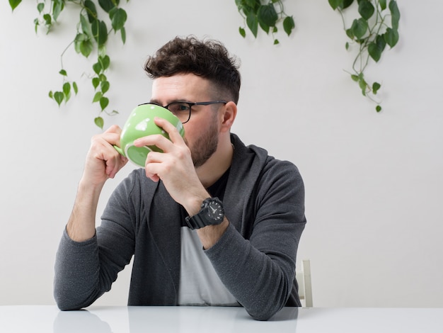 jeune homme assis dans des lunettes montres veste grise buvant son jus de la tasse verte avec plante sur blanc