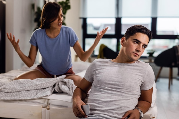 Photo gratuite jeune homme assis près du lit et ignorant sa petite amie qui se dispute avec lui