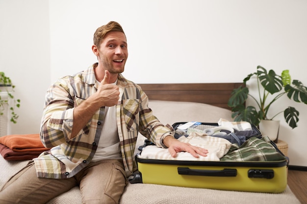 Photo gratuite jeune homme attrayant souriant montrant les pouces en l'air montrant ses vêtements à l'intérieur de la valise se prépare pour