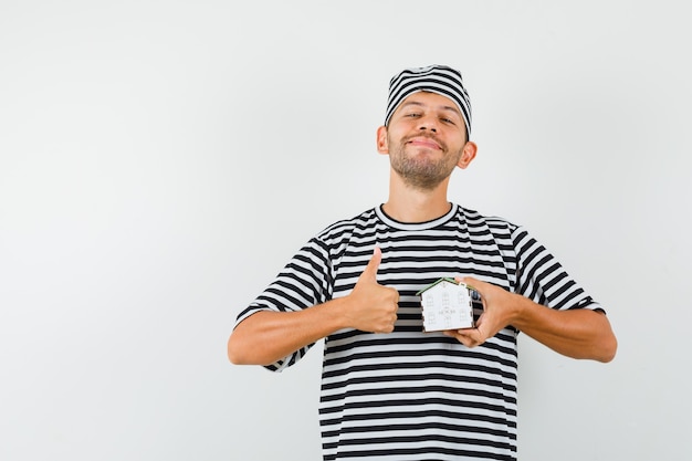 Jeune homme au chapeau t-shirt rayé tenant le modèle de maison montrant le pouce vers le haut et à la confiance