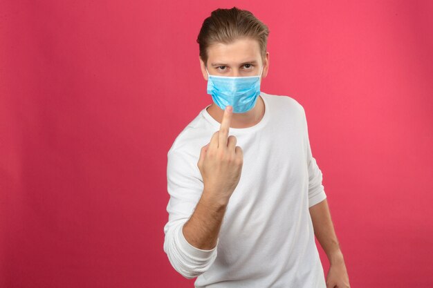 Jeune homme au masque de protection médicale montrant l'expression grossière du majeur sur fond rose isolé