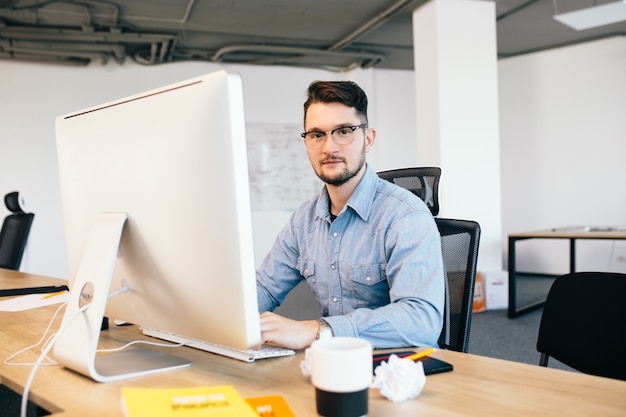Jeune homme aux cheveux noirs en glasess et une chemise bleue travaille avec un ordinateur sur son bureau au bureau. Il sourit à la caméra.