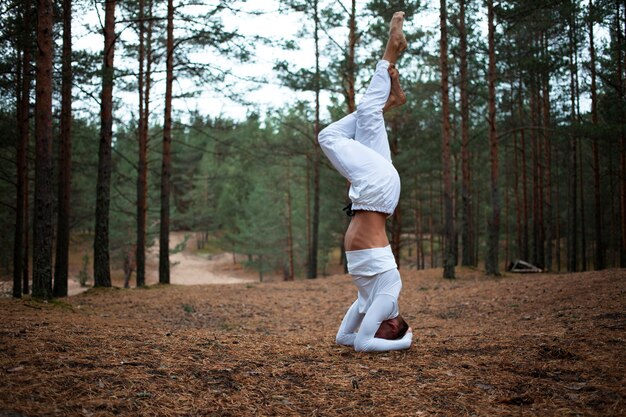 Jeune homme aux pieds nus en vêtements blancs faisant une variation de la position de yoga Salamba Shirshasana sur le sol dans la forêt, croisant les jambes. Prise de vue en extérieur d'une formation de yogi avancée dans les bois, en équilibre sur les mains