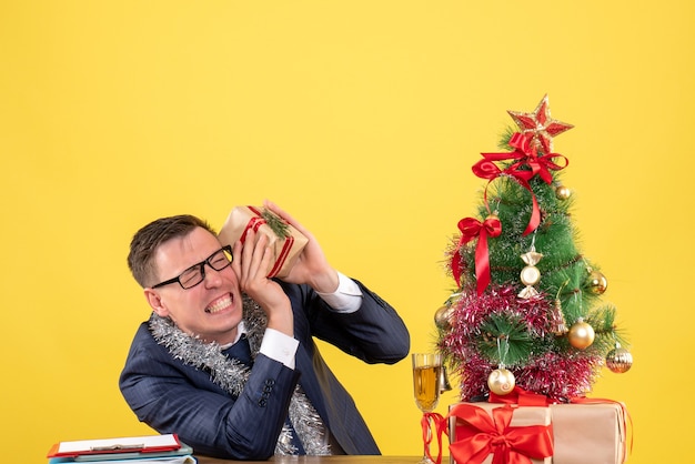 Jeune homme aux yeux fermés tenant un cadeau assis à la table près de l'arbre de Noël et présente sur jaune