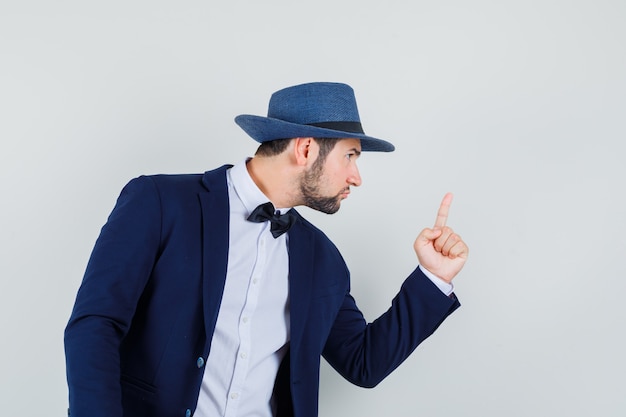 Jeune homme avertissant quelqu'un avec le doigt en costume, chapeau et à la vue de face, nerveux.