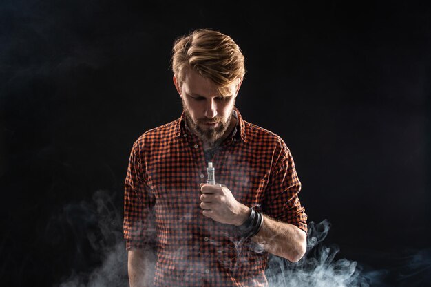 Un jeune homme avec une barbe et une coiffure élégante en chemise, fumant une cigarette, une vipère, une chambre, un studio, de la fumée, du plaisir. Fond noir