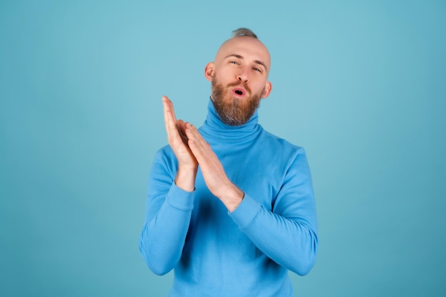 Un jeune homme avec une barbe rouge dans un col roulé sur fond bleu applaudit joyeusement, accueillant