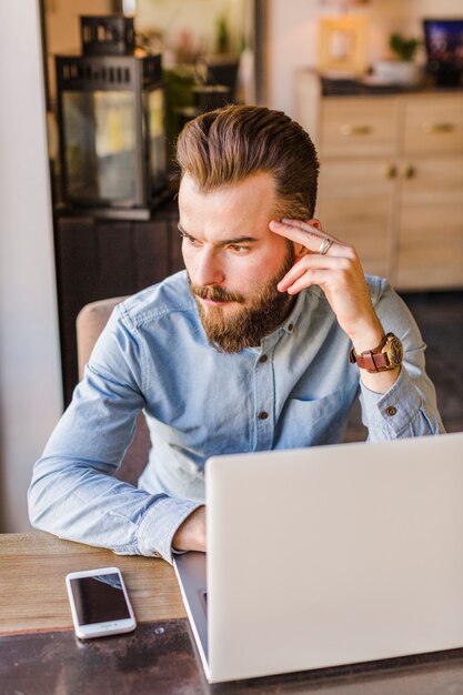 Jeune homme barbu assis au restaurant avec ordinateur portable et téléphone portable sur le bureau