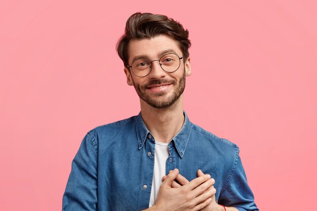 Un jeune homme barbu au cœur bienveillant garde les deux paumes sur la poitrine, regarde positivement, exprime des sentiments véridiques