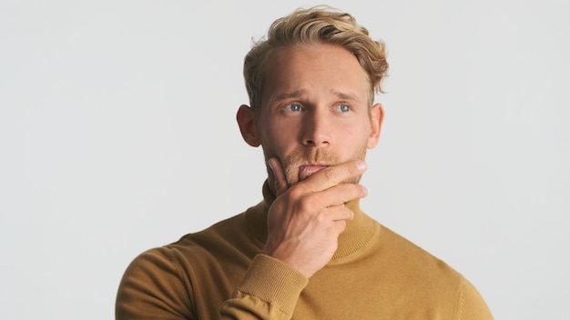 Photo gratuite jeune homme barbu blond attrayant garder la main sur le menton en pensant à la caméra isolé sur fond blanc