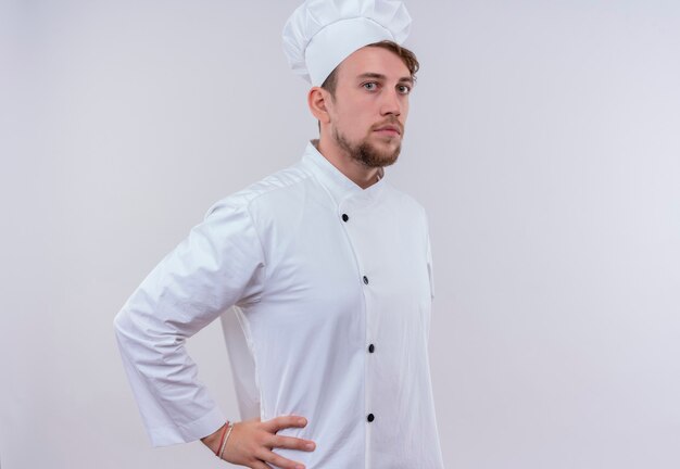 Un jeune homme barbu chef sérieux en uniforme blanc tenant la main sur sa taille tout en regardant sur un mur blanc