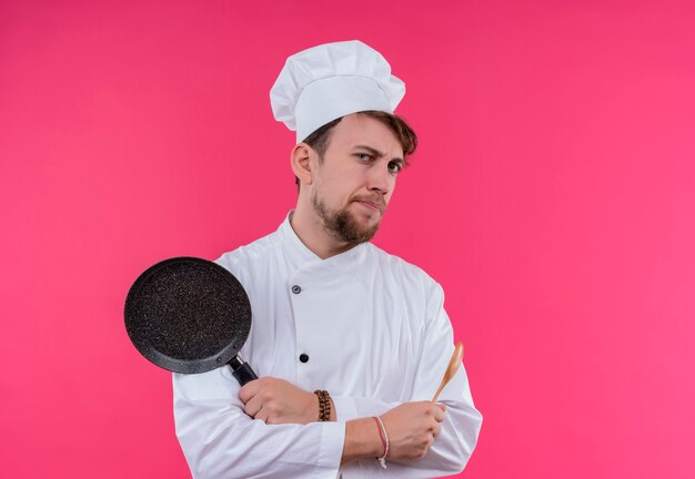 Un jeune homme barbu chef sérieux en uniforme blanc tenant une poêle avec une cuillère en bois tout en regardant sur un mur rose