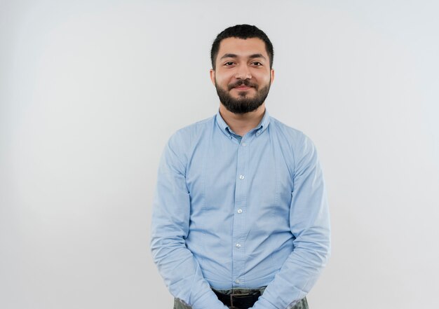 Jeune homme barbu en chemise bleue à l'avant souriant confiant debout sur un mur blanc