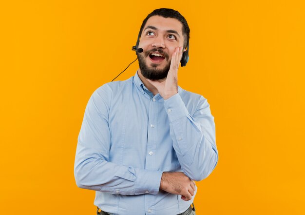 Jeune homme barbu en chemise bleue avec des écouteurs avec microphone appelant quelqu'un avec la main près de la bouche en souriant