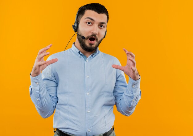 Jeune homme barbu en chemise bleue avec des écouteurs avec microphone montrant un geste de grande taille avec les mains surpris