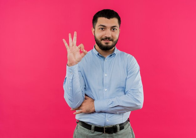 Jeune homme barbu en chemise bleue montrant signe ok souriant