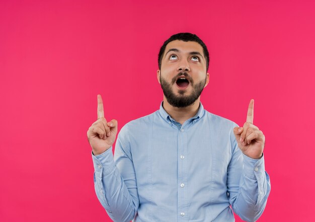 Jeune homme barbu en chemise bleue à la recherche de l'index pointant heureux et excité vers le haut