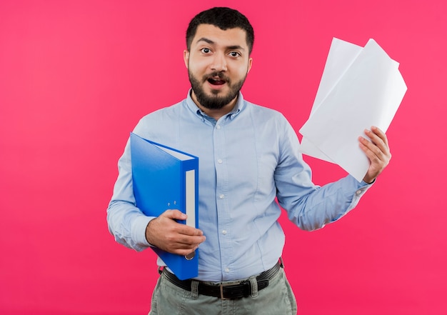 Jeune homme barbu en chemise bleue tenant le dossier et les pages blanches heureux et excité