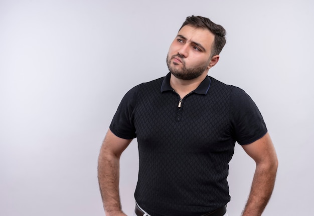 Photo gratuite jeune homme barbu en chemise noire à côté avec une expression pensive pensant, ayant des doutes