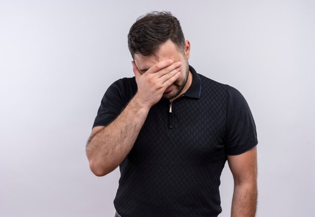 Jeune homme barbu en chemise noire à la fatigue et l'ennui couvrant les yeux avec bras