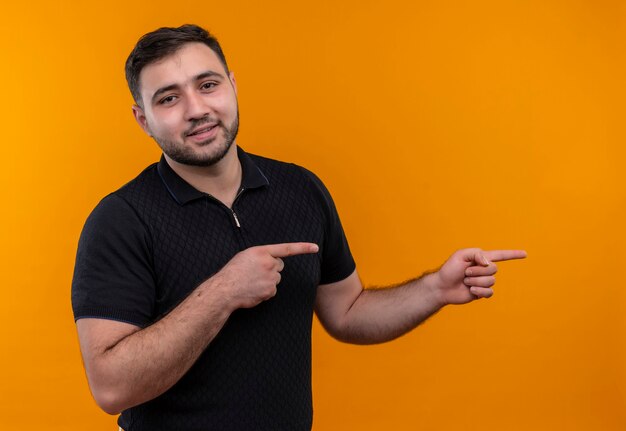 Jeune homme barbu en chemise noire pointant avec l'index sur le côté regardant la caméra avec une expression confiante