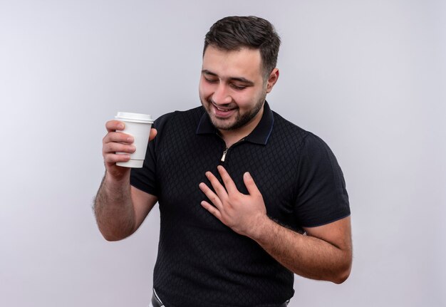 Jeune homme barbu en chemise noire tenant une casquette de café souriant positif et heureux