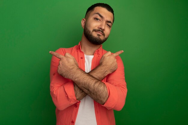 Jeune homme barbu en chemise orange avec une expression confiante pointant avec index sur les côtés opposés debout sur mur vert