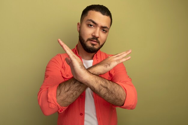 Photo gratuite jeune homme barbu en chemise orange faisant le geste d'arrêt croisant les mains avec un visage sérieux debout sur un mur léger