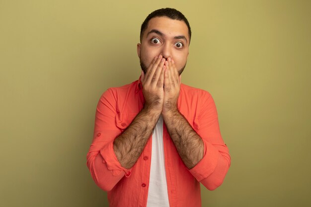 Jeune homme barbu en chemise orange surpris couvrant la bouche avec les mains debout sur un mur léger