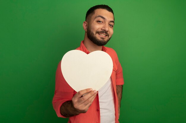 Jeune homme barbu en chemise orange tenant coeur en carton à la recherche avec le sourire sur le visage debout sur le mur vert