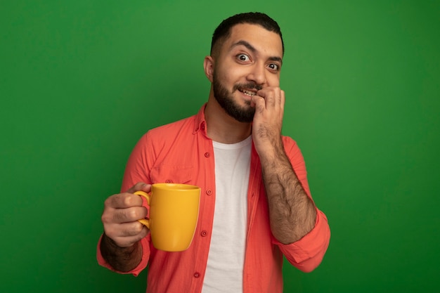 Jeune homme barbu en chemise orange tenant une tasse a souligné et nerveux mordant les ongles debout sur le mur vert