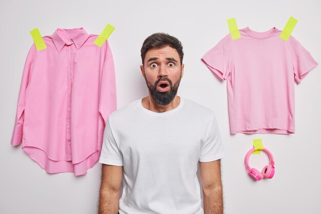 Un jeune homme barbu choqué regarde avec une expression étonnée effrayée porte un t-shirt décontracté pose sur fond blanc des vêtements roses et des écouteurs collés au mur réagit aux rumeurs inattendues