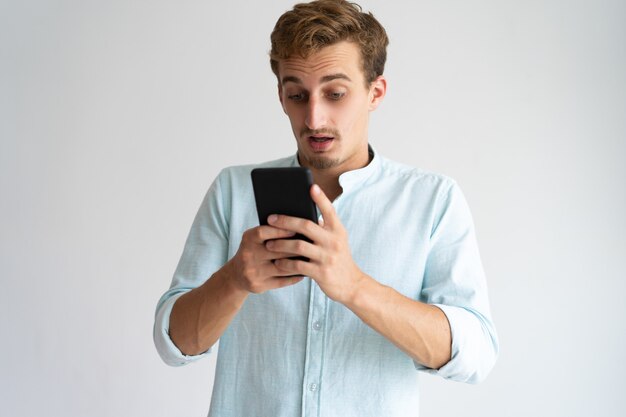 Jeune homme barbu choqué en train de lire les nouvelles d&#39;Internet sur un smartphone.